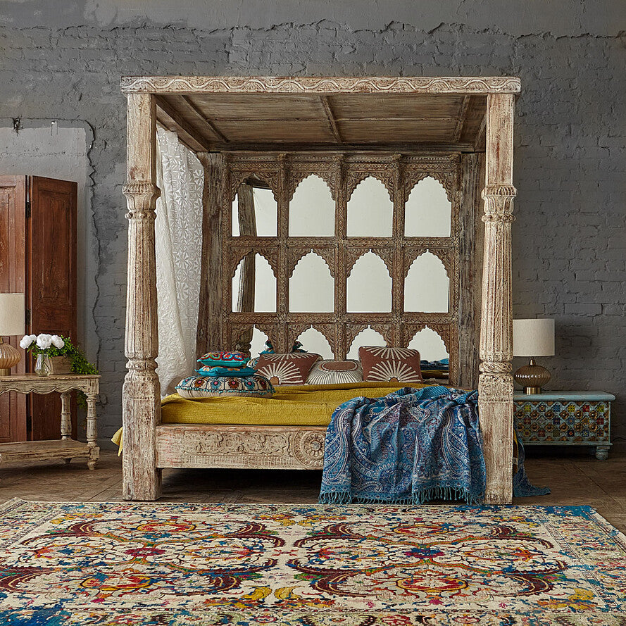 Wooden four-poster bed with intricate designs in a room with a textured gray wall and colorful rug.