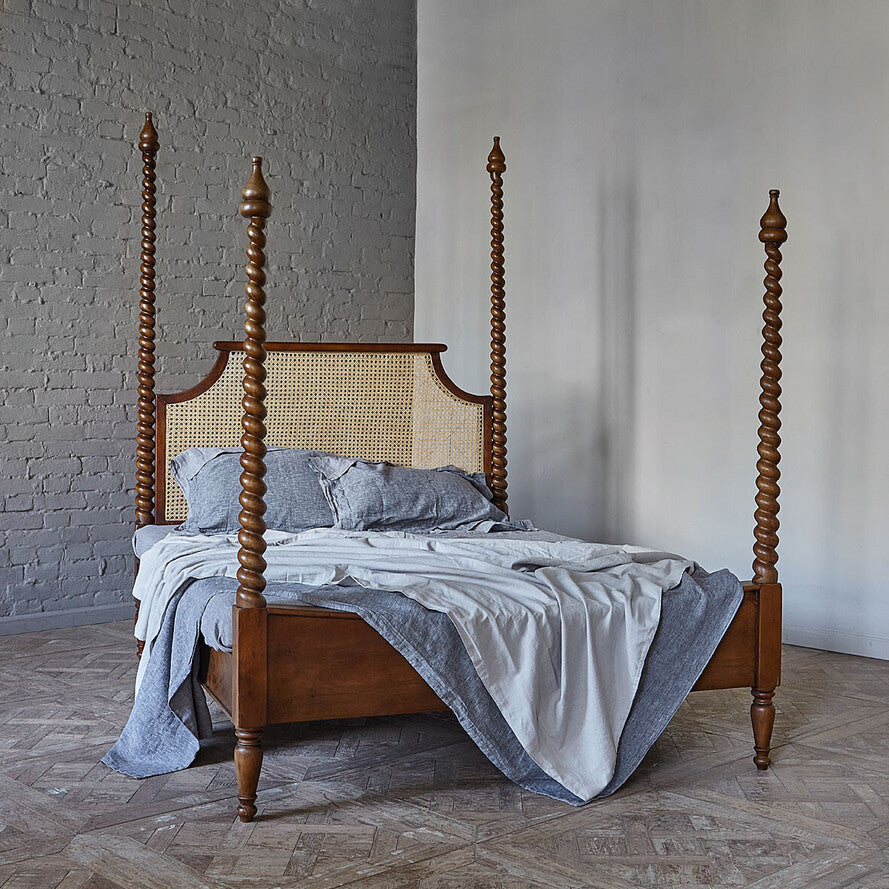 Wooden four-poster bed with wicker headboard in a room with gray walls and wooden floor.