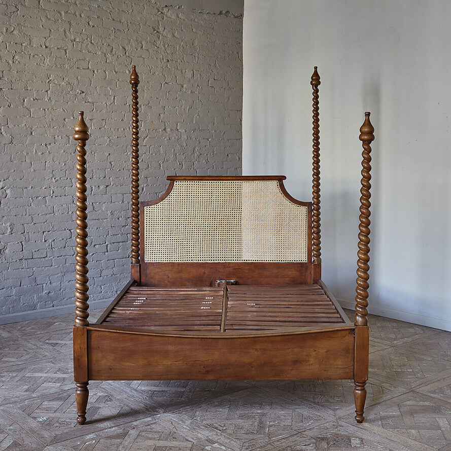 Wooden four-poster bed with a woven headboard in a room with a textured wall and wooden floor.
