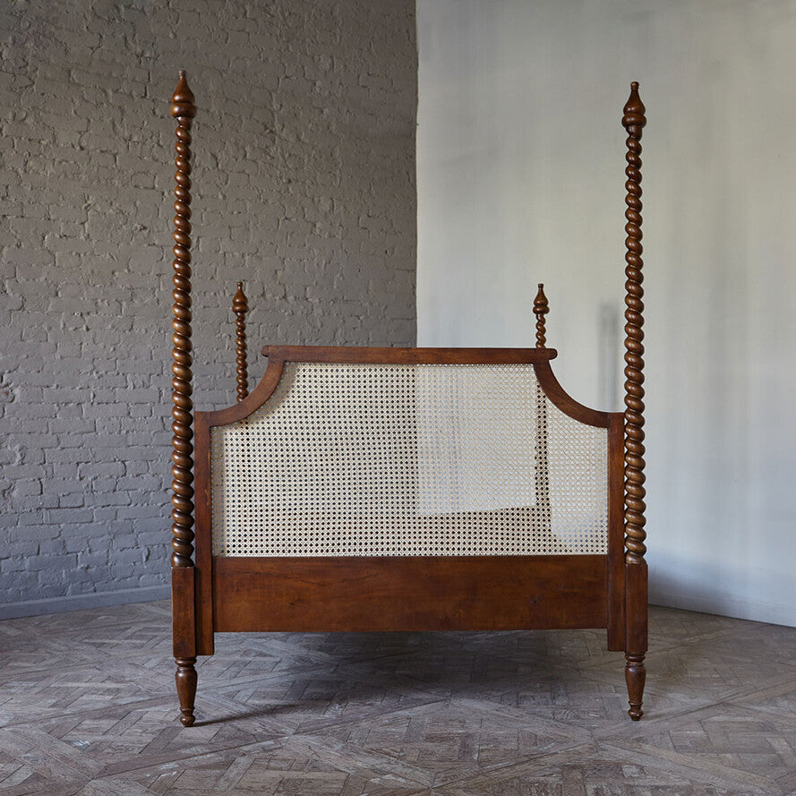 Wooden four-poster bed with a cane backrest against a gray wall.