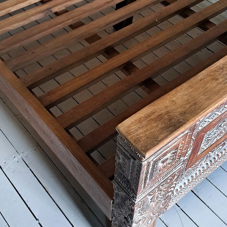 Wooden bed frame with intricate carvings on a light wooden floor.